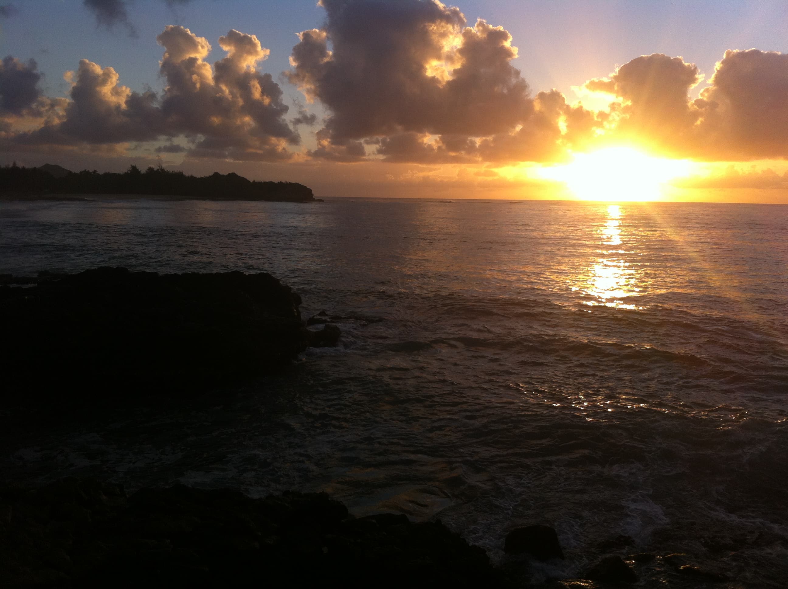 A dramatic tropical sunrise breaks through stormy clouds over rugged coastline, casting golden light across churning waters.
